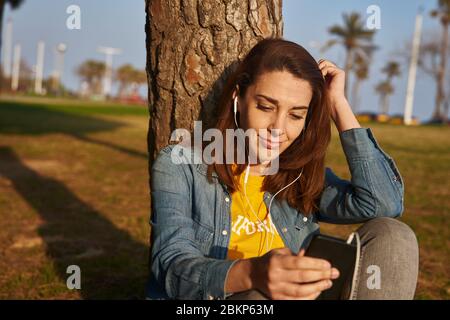 Une femme assise contre un tronc d'arbre à écouter quelque chose par des écouteurs et un téléphone portable. Banque D'Images