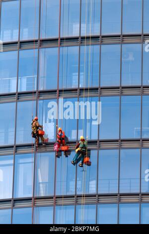 Trois hommes travaillent en vêtements de travail rouges et sombres pour nettoyer les fenêtres extérieures d'un gratte-ciel d'affaires. Extérieur Banque D'Images