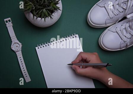 Les baskets blanches, les montres, un pot avec succulent et une main de womans écrit dans un carnet avec un stylo sur le fond vert. Banque D'Images