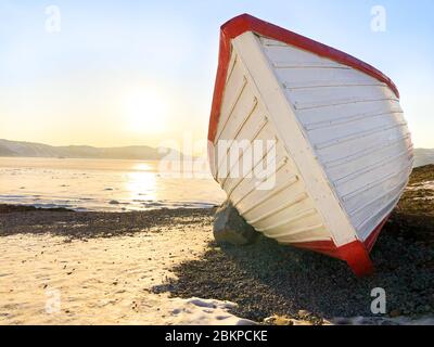 Photo d'un bateau en bois sur le bord de mer de Magadan, Russie. Bateau blanc à rayures rouges. Coucher de soleil sur l'arrière-plan. Ciel bleu clair et glace sur la mer en hiver Banque D'Images