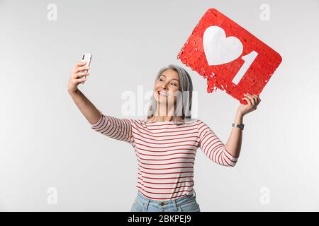 Image d'une femme adulte mature avec de longs cheveux blancs tenant un téléphone cellulaire et un symbole de coeur sur l'étiquette isolée sur fond gris Banque D'Images