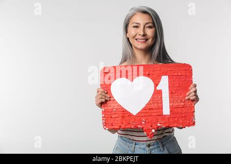 Image d'une femme adulte mûre heureuse avec de longs cheveux blancs tenant le symbole du cœur sur l'étiquette isolée sur fond gris Banque D'Images
