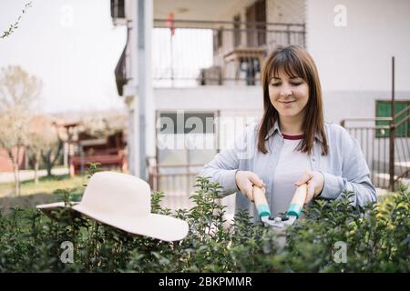 Jeune femme façonnant des arbustes de jardin avec un outil de coupe. Femme souriante coupant l'usine en utilisant des ciseaux de jardin tout en se tenant devant une maison. Banque D'Images