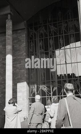 1961, photo historique des personnes devant l'entrée des vitraux de la nouvelle cathédrale, Coventry, Angleterre. Lourdement bombardé pendant la période éclair de la Seconde Guerre mondiale, en mai 1962, un nouveau bâtiment « moderniste » a été officiellement ouvert sur le site des ruines de l'ancienne cathédrale. Le nouveau bâtiment a été conçu par Basil Spence dans le style architectural radical 'Brutaliste', commun à cette époque. Banque D'Images