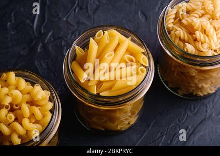 Trois pots en verre de suite avec variété de pâtes de blé doré non cuites sur fond noir foncé texturé, vue en angle macro Banque D'Images