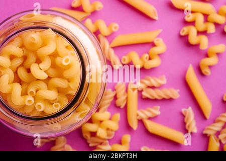 Un pot en verre avec une variété de pâtes de blé doré non cuites sur fond rose minimal, vue de dessus macro Banque D'Images