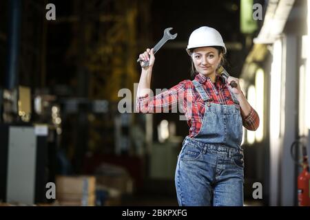 Jeune fille dans une robe de travail et chapeau de dur blanc tenant une grosse clé et hummer dans une usine. Femme en uniforme de travail. Processus de travail. Banque D'Images