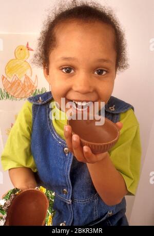 BÉBÉ DE PÂQUES Africain américain Carribbean fille de 4 ans piquant dans une coquille d'oeuf de Pâques au chocolat, avec sa poussette de Pâques dessinant sur son mur derrière Banque D'Images