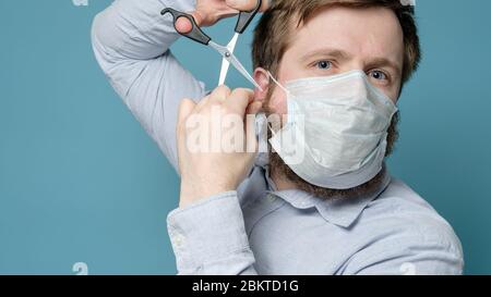 L'homme essaie de couper sa barbe tiraillée avec des ciseaux car le masque médical ne tient pas bien. Concept de sécurité. Pandémie. Banque D'Images