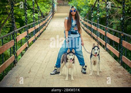 Jolie fille et ses animaux de compagnie. La femme de rapaces de bagarles avec ses chiens husky a isolé le parc vert en restant sur le pont les trois regardant votre caméra. Tak Banque D'Images