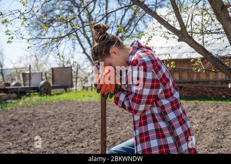 Une fille brunette fatiguée en gants de travail orange penchée sur une pelle, se reposant tout en travaillant dur dans le jardin Banque D'Images