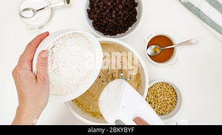 Recette de biscuits aux pépites de chocolat. Chef ajoute de la farine dans une pâte à biscuits, gros plan sur fond blanc, vue d'en haut Banque D'Images