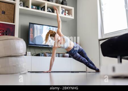 Jolie femme sportive travaillant à la maison, faisant des pilates exercice devant la télévision dans son salon. Distanciation sociale. Restez en bonne santé et Banque D'Images