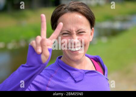 Mignonne amusante femme aimante donnant un V-sign avec un sourire éclatant alors qu'elle prend une pause de jogging dehors dans la campagne du printemps Banque D'Images