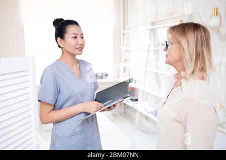 Cosmetologiste asiatique amical avec presse-papiers fournissant au client des informations sur la procédure dans le salon de beauté Banque D'Images