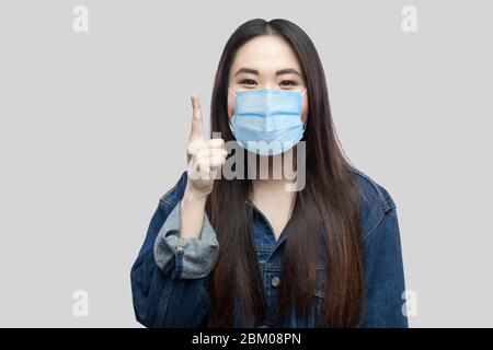 Idée ! Portrait de la belle fille asiatique de brunette excimée avec masque médical chirurgical en bleu veste denim debout et regardant l'appareil photo, doigt vers le haut. Dans Banque D'Images