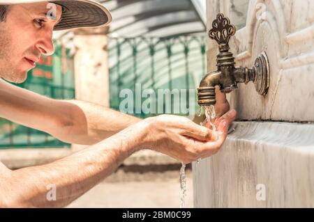 Un jeune homme boit de l'eau avec sa main d'une ancienne fontaine turque sur un mur de marbre. Eau courante du robinet. Banque D'Images