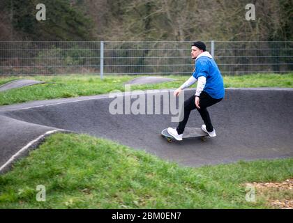 Vue latérale d'un jeune homme concentré dans des vêtements de sport chauds et chapeau sur un skateboard le long d'un chemin d'asphalte vallonné, effectuant des exercices d'équilibre dans le parc de skate Banque D'Images