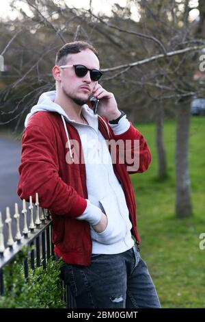 Homme tendance et chic, vêt de vêtements décontractés et de lunettes de soleil, en conversation téléphonique, debout dans la poche et en se penchant sur la clôture du parc de la ville Banque D'Images