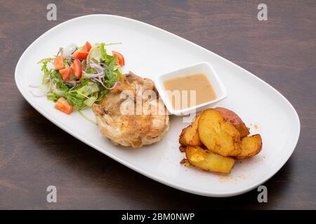 Poulet rôti avec sauce, pommes de terre cuites au four et salade mixte dans une assiette en céramique blanche sur une table en bois Banque D'Images