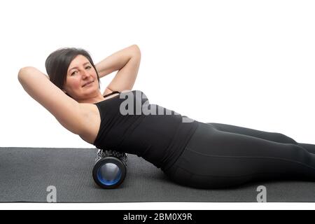 Une femme d'âge moyen avec une peau sagy sur un tapis de gymnastique avec rouleau myofascial fait un exercice sur son dos sur un fond blanc. Banque D'Images