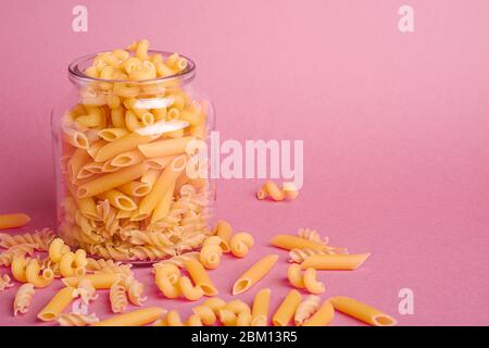 Un pot en verre avec une variété de pâtes de blé doré non cuites sur fond rose minimal, vue en angle macro, espace de copie Banque D'Images