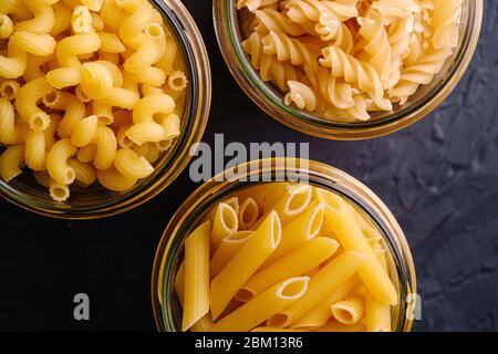 Trois pots en verre avec variété de pâtes de blé doré non cuites sur fond noir foncé texturé, vue de dessus macro Banque D'Images