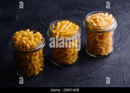 Trois pots en verre de suite avec variété de pâtes de blé doré non cuites sur fond noir foncé texturé, vue en angle Banque D'Images