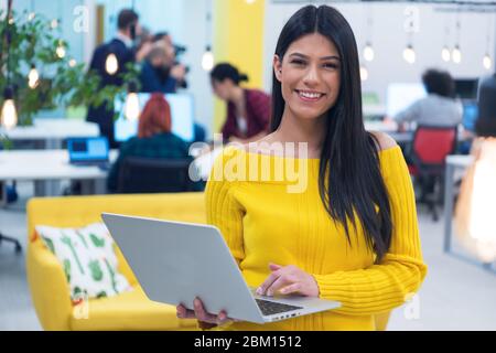 Portrait d'une jeune femme d'affaires décontractée et heureuse debout avec un ordinateur portable sur son lieu de travail dans un espace de travail moderne. Banque D'Images