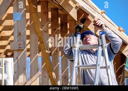 Un constructeur à poil gris recueille le cadre d'une maison de campagne en bois avec un tournevis sans fil le jour du printemps contre un ciel bleu. Le a physique Banque D'Images