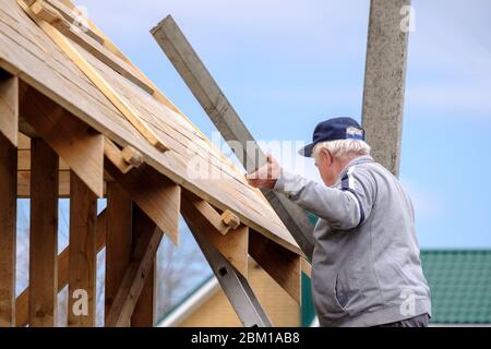 Un constructeur à poil gris recueille le cadre d'une maison de campagne en bois debout sur les escaliers contre le ciel bleu. L'activité physique de l'elde Banque D'Images