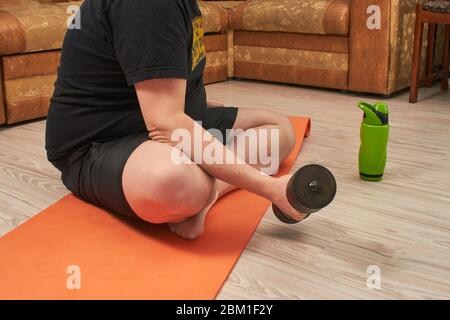 fat homme caucasien assis en position lotus sur un tapis de fitness avec des haltères à la maison en quarantaine Banque D'Images