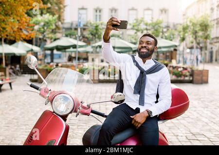 Jeune homme souriant et sombre, avec chemise blanche et pantalon noir, assis sur le scooter rouge moderne et faisant des selfies photo sur le fond de Banque D'Images