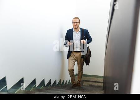 Image de beau heureux positif jeune homme d'affaires à l'intérieur marchant à l'étage en utilisant le téléphone mobile. Banque D'Images