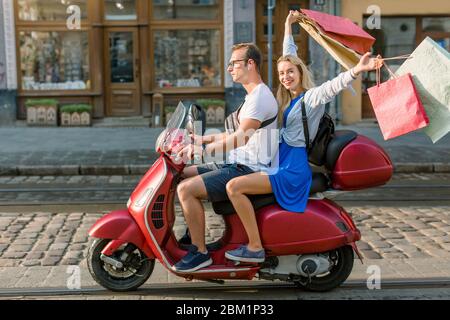 L'été, le style de vie urbain. Jeune couple tendance s'amusant sur un scooter rouge vintage dans les rues, jolie femme blonde souriante a ses sacs de shopping Banque D'Images