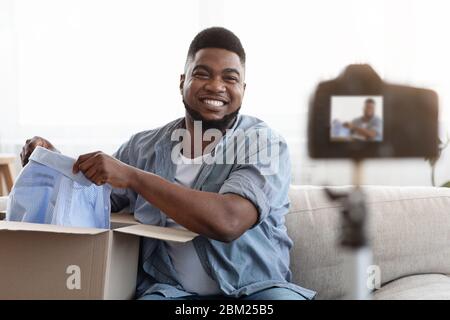 Déballage du Vlog. Sourire Noir Homme déballage boîte avec de nouveaux vêtements à la caméra Banque D'Images