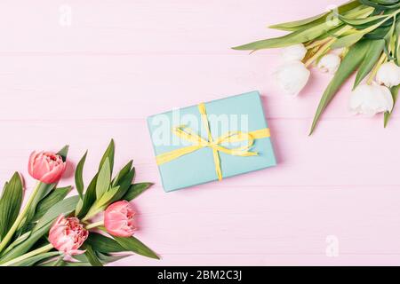 Boîte cadeau à côté de bouquets de fleurs de tulipe fraîches et de feuilles vertes sur fond rose, composition de la pose plate. Banque D'Images