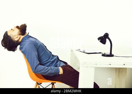 Homme barbu, barbe longue. Hipster caucasien brutal et non rasé, avec moustache assise sur une chaise orange près d'une table en bois blanc avec bloc-notes ouvert et lampe noire dessus, isolé sur fond blanc Banque D'Images