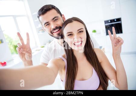 Portrait de positif gai couple marié homme femme séjour quarantaine maison faire selfie montrer v-signe blogging dans la maison de cuisine à l'intérieur Banque D'Images