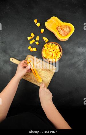 Une femme coupe une citrouille sur une planche à découper en bois sur une table en pierre. Mains de femmes avec couteau en acier, morceaux de courge de noyer cendré dans un bol en bois sur fond noir Banque D'Images