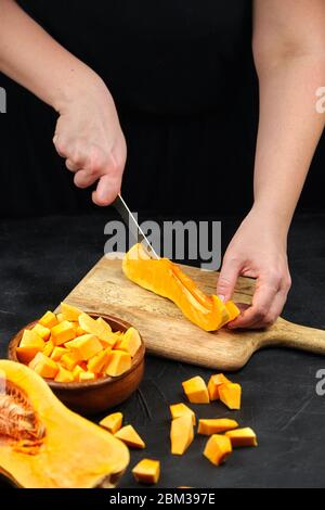 Une femme coupe une citrouille sur une planche à découper en bois sur une table en pierre. Mains de femmes avec couteau en acier, morceaux de courge de noyer cendré dans un bol en bois sur fond noir Banque D'Images