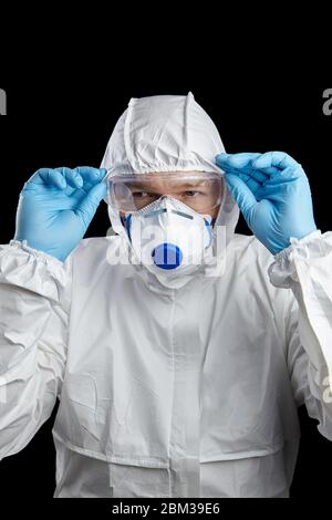 homme en costume de protection blanc et masque respiratoire, gants et lunettes bleus jetables, isolés sur fond noir Banque D'Images