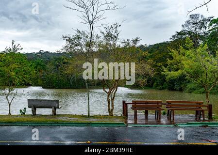 Par temps pluvieux, trois bancs vides font face à un lac, entouré de bois. Des nuages et une partie de l'asphalte humide sont également vus ici. Banque D'Images