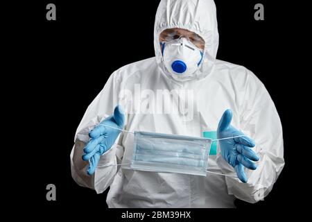 homme en costume de protection blanc et masque respiratoire avec masque médical dans les mains, gants et friandises bleus jetables, isolés sur fond noir Banque D'Images