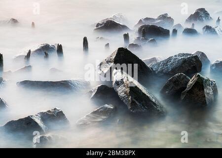 Côte de mer avec des vagues qui s'écrasant sur les rochers. Exposition longue, image floue de l'eau, la nébuleuse se déforme entre les roches du rivage. Le ciel couvert gris. Banque D'Images