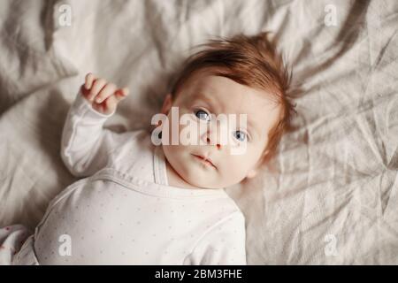 Portrait en gros plan de bébé casien mignon nouveau-né. Adorable enfant drôle avec des yeux gris bleu et des cheveux rouges couché sur le lit regardant la caméra. Banque D'Images