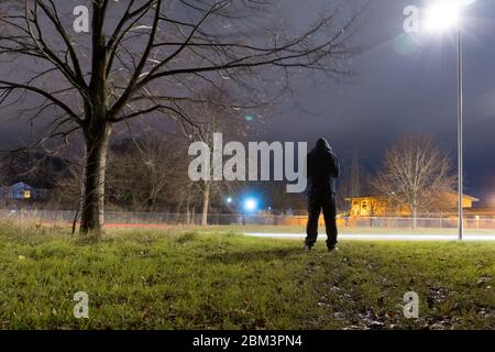 Une silhouette à capuche, silhouetée contre une lumière de rue sur le bord de la ville la nuit Banque D'Images