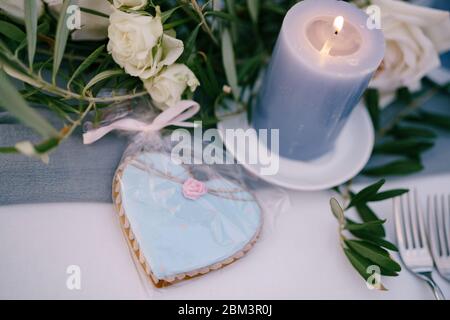 Biscuit en forme de coeur bleu en forme de pain d'épice dans un paquet cadeau se trouve sur la table, près d'une bougie de cire épaisse brûlante dans des fleurs et des branches d'olive Banque D'Images