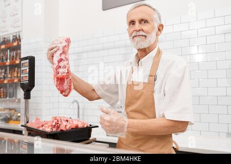 Foyer sélectif de boucher mâle senior tenant un morceau de viande fraîche brute rouge en main et montrant le pouce vers le haut. Homme joyeux derrière le comptoir du magasin montrant de la viande, bol du réfrigérateur sur des balances. Banque D'Images