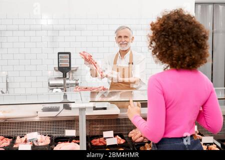 Foyer sélectif de boucher masculin senior tenant un morceau de viande fraîche brute rouge et montrant à la femme avec des cheveux bouclés. Homme joyeux derrière le comptoir du magasin montrant de la viande, bol du réfrigérateur sur des balances. Banque D'Images